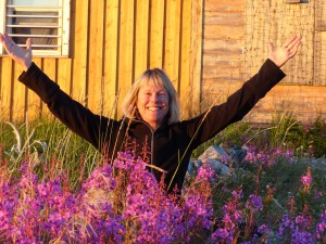 Gutsy Traveler: A woman, like travel expert Marybeth Bond, stands among purple flowers, raising her arms and smiling in front of a wooden building in bright sunlight.