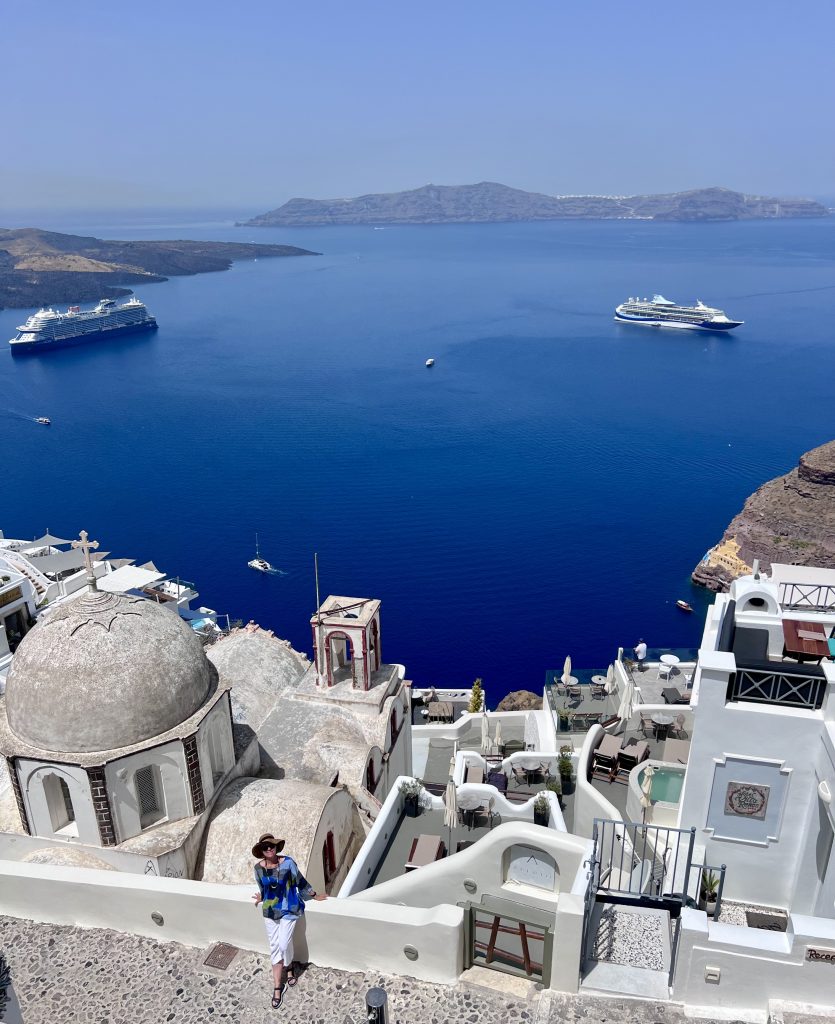 Gutsy Traveler: A coastal view with white buildings in the foreground, a dome-shaped structure, and two cruise ships set for Mediterranean cruises in the blue water of the sea against a backdrop of distant mountainous islands.