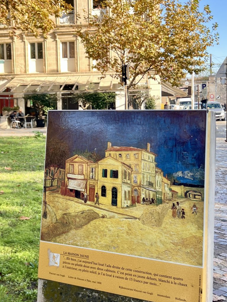 Gutsy Traveler: A historical marker with a painting of a yellow house stands in a sunny park, surrounded by grass and trees, evoking the charming ambiance experienced on a France river cruise for 50+ travelers, with a café and buildings in the background.
