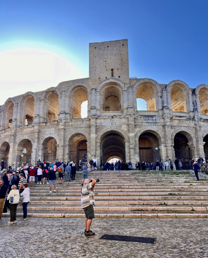 Gutsy Traveler: Amidst a sunlit day, a traveler from a France River Cruise for adventurers aged 50+ snaps a photo in front of an ancient stone amphitheater, where the steps buzz with curious onlookers basking under the clear blue sky.