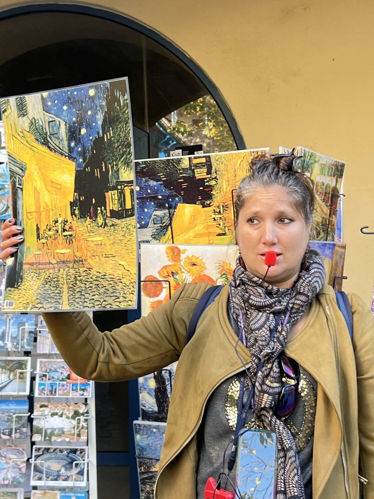 Gutsy Traveler: A person holding up Van Gogh art prints, wearing a green jacket and patterned scarf, stands in front of an outdoor display—a perfect snapshot for those enjoying a France River Cruise for travelers 50+.
