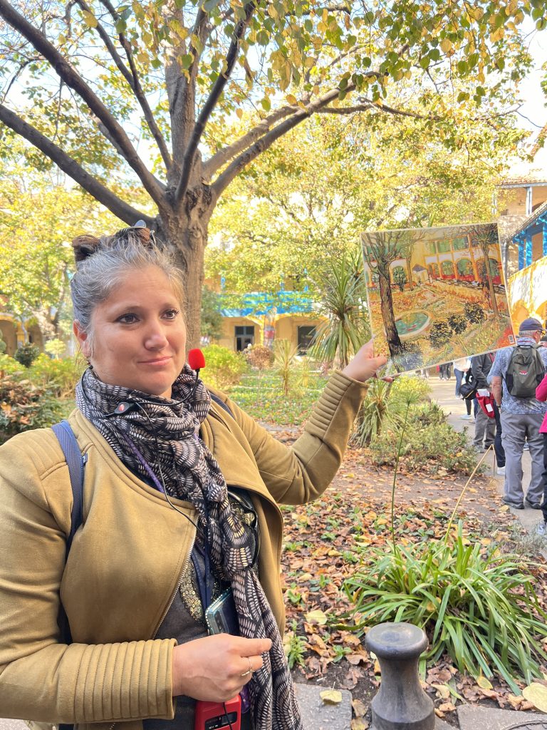 Gutsy Traveler: Amidst the rustling leaves, a woman holds up a colorful painting, her scarf fluttering in the breeze. It's like capturing the serene beauty of a France river cruise for travelers 50+, blending art with nature's tranquility.