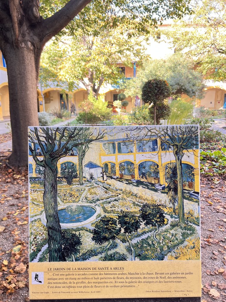 Gutsy Traveler: A painting of a garden, reminiscent of scenes along a France river cruise for travelers 50+, is displayed in front of a real garden setting, with trees and a building in the background. The exhibit is titled "Le Jardin de la Maison de Santé à Arles.