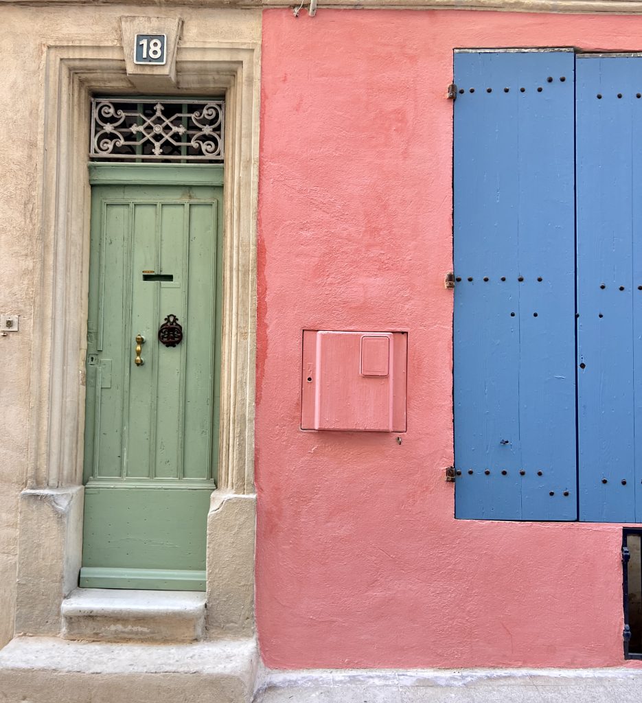 Gutsy Traveler: A green door with a decorative grate and number 18 on the left, surrounded by beige, welcomes those embarking on a France River Cruise for travelers 50+. Adjacent are pink walls with a matching mailbox and closed blue shutters on the right, echoing the charm of scenic French streets.