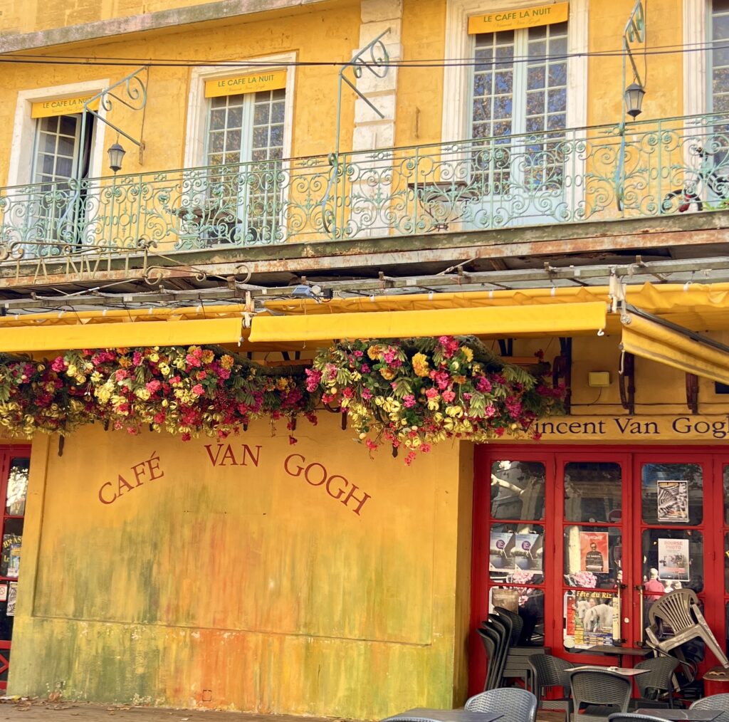 Gutsy Traveler: The yellow building, marked "Café Van Gogh," features red doors and windows adorned with floral decorations, capturing the essence of a French getaway. Outside, tables and chairs await patrons who might stop by during their Van Gogh art tour or river cruise adventure.