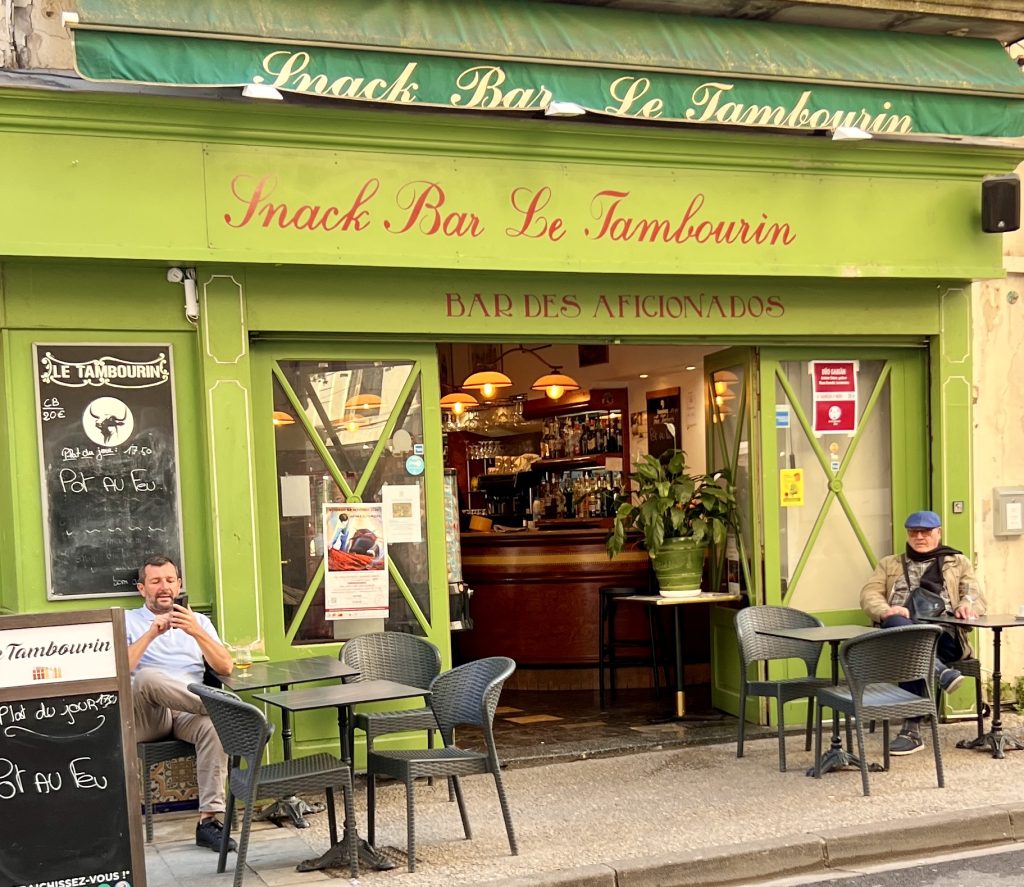 Gutsy Traveler: A green snack bar with outdoor seating, reminiscent of a stop along a France River Cruise for Travelers 50+. Two men sit at tables, enjoying the relaxed ambiance, as the charming interior is visible through the open doors.