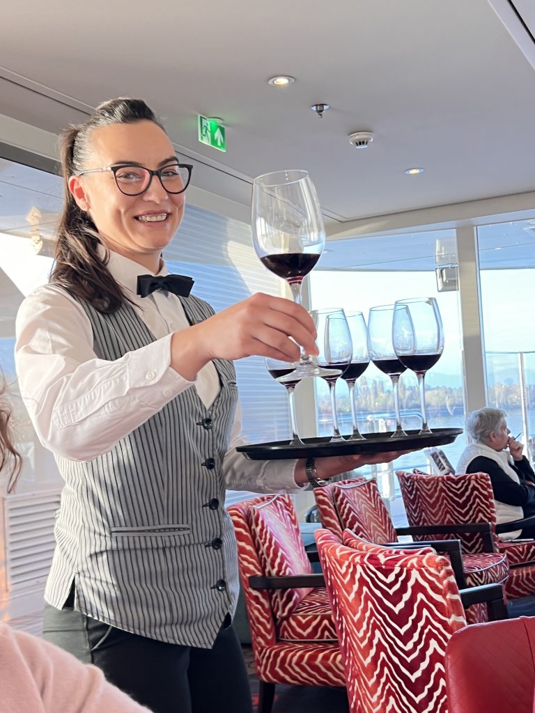 Gutsy Traveler: A server with glasses and a bow tie gracefully holds a tray of red wine glasses in a restaurant with red-patterned chairs, evoking the elegance of a France river cruise for travelers 50+.