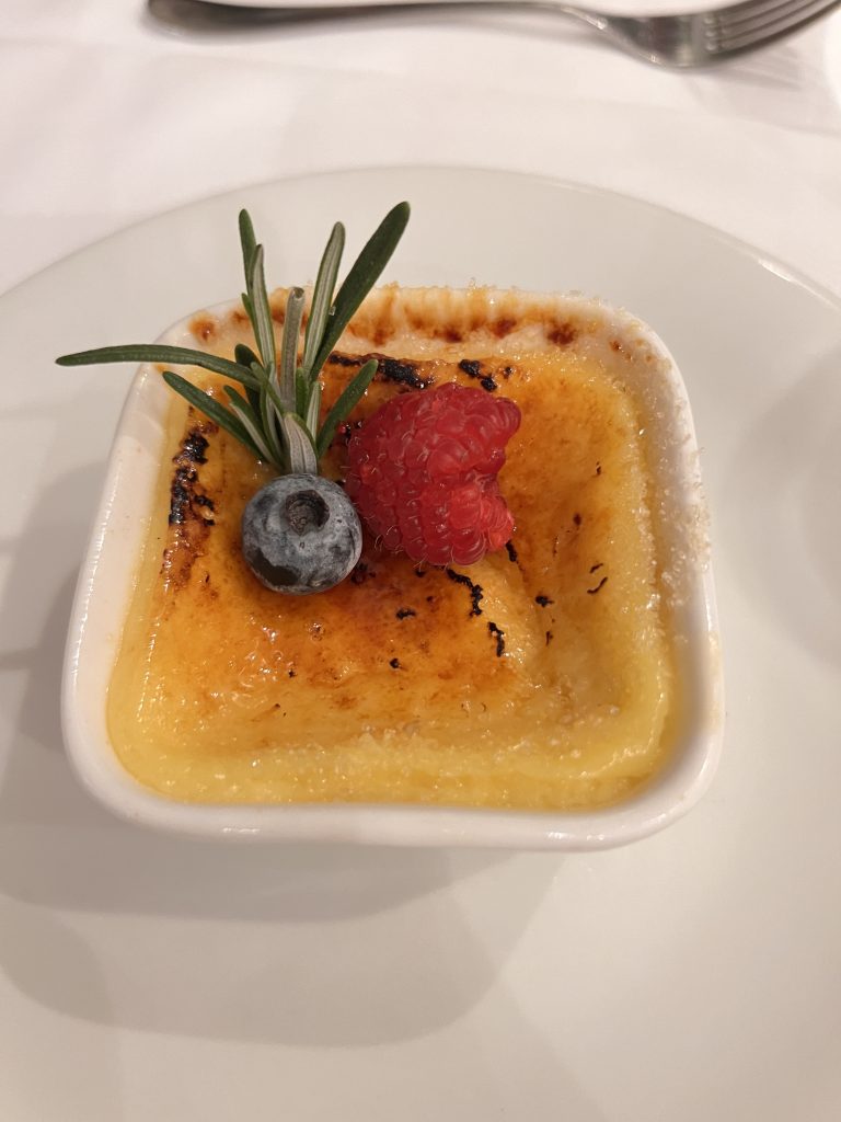 Gutsy Traveler: A small crème brûlée in a square dish, graced with a raspberry, blueberry, and a sprig of rosemary, evokes the charm of a culinary experience on a France River Cruise for travelers 50+, elegantly presented on a white plate.