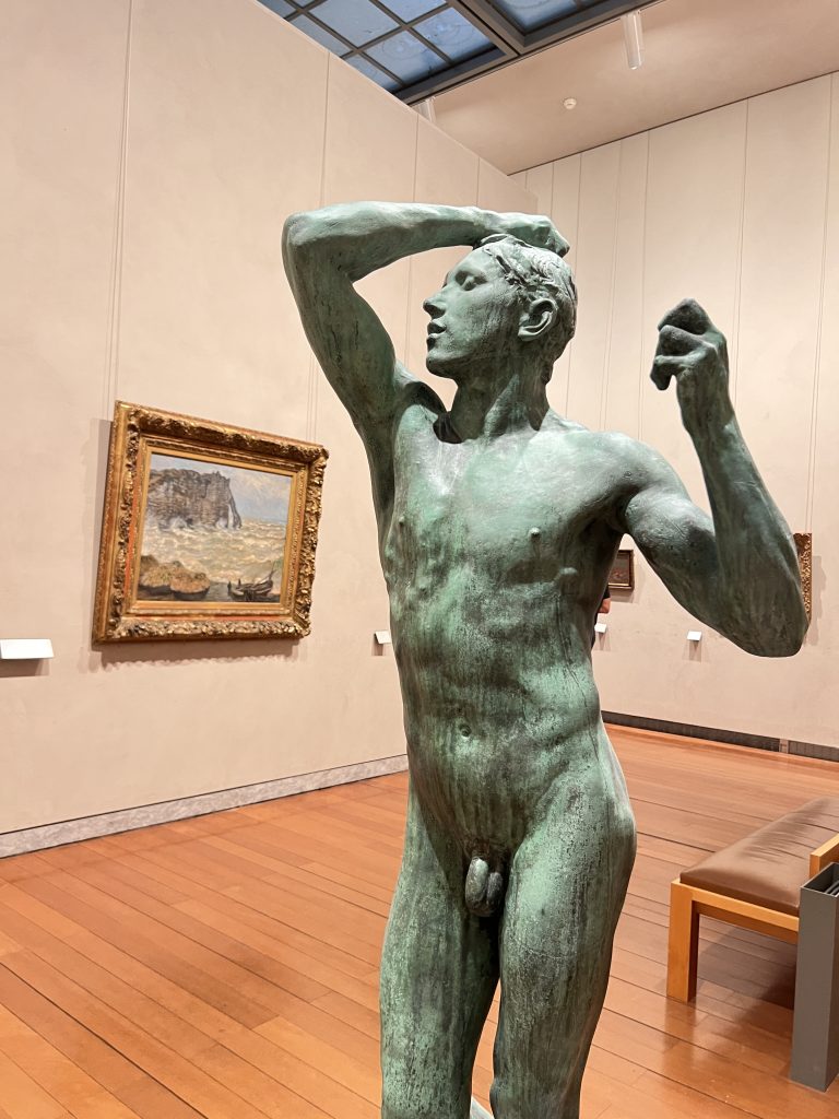 Gutsy Traveler: In a museum gallery, a bronze statue of a standing nude male figure stands prominently amid exquisite paintings, offering an unexpected cultural detour reminiscent of treasures discovered on a France River Cruise for travelers 50 and above.