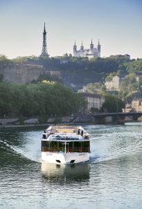 Gutsy Traveler: A riverboat cruises on a calm river, offering travelers 50+ a stress-free travel experience with views of a hilltop basilica and tower in the background. Lush greenery and city buildings line the riverbank, enhancing this enchanting France river cruise.