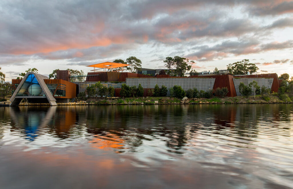 Gutsy Traveler: A modern building with angular architecture sits by a calm river, surrounded by trees and under a partly cloudy sky at sunset—a true treasure hunt for lovers of must-see museums on vacation.
