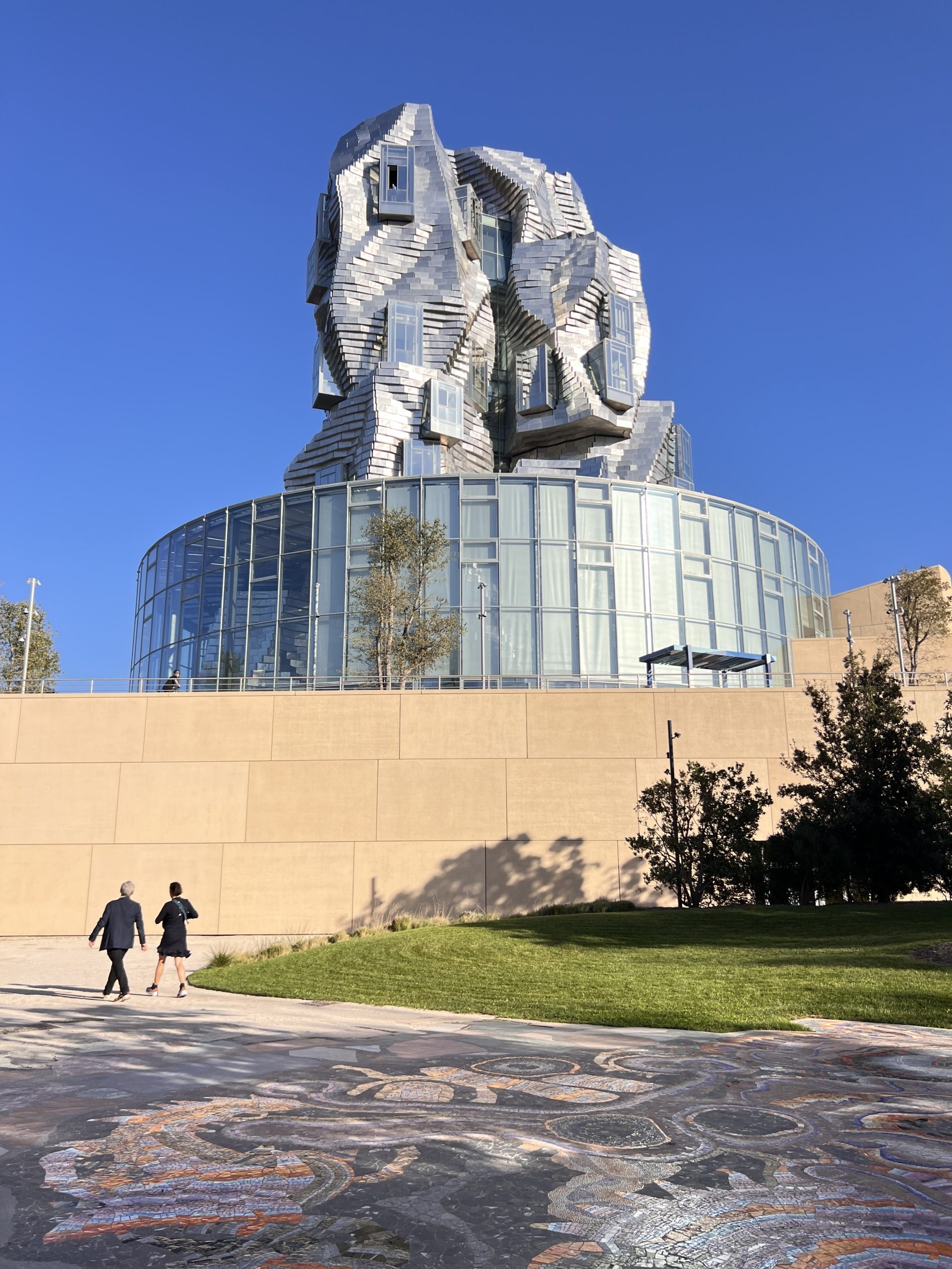 Gutsy Traveler: Two people walk past a modern building with a round glass base and an irregular, metallic upper structure—a must-see museum for anyone on vacation under a clear blue sky.
