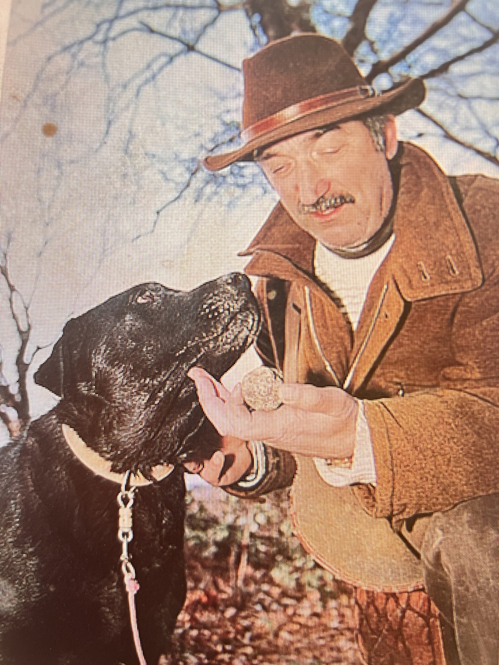 Gutsy Traveler: A man in a brown jacket and hat shares a truffle with a black dog on a leash outdoors, showcasing an authentic truffle hunting experience amid leafless trees, reminiscent of Provence, France.