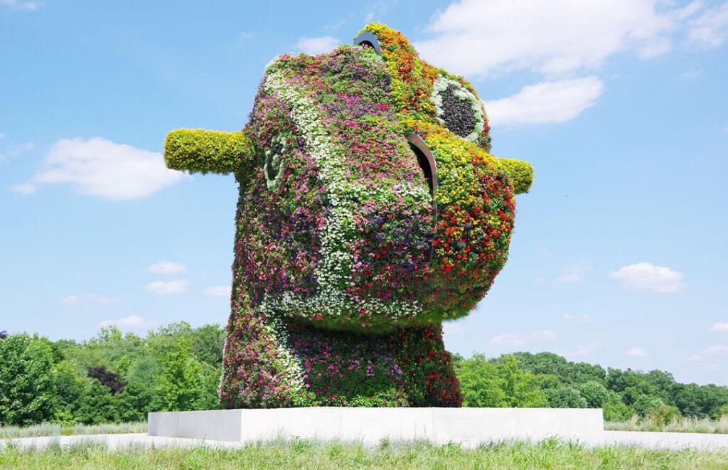 Gutsy Traveler: A large outdoor sculpture of an animal head, adorned with colorful flowers and greenery, stands on a pedestal in a grassy field—like a hidden gem for any vacation treasure hunt beneath the blue sky and framed by trees.