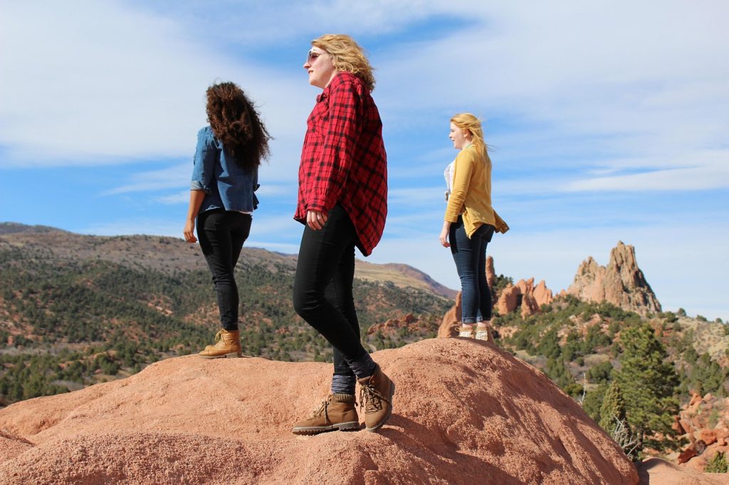 Gutsy Traveler: Three women stand on a rocky hill in Sedona, overlooking vortex sites, with trees and distant mountains beneath a partly cloudy sky.