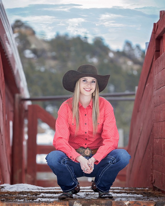 Gutsy Traveler: A woman wearing a black cowboy hat, red shirt, jeans, and boots squats on a wooden walkway with Sedona trees behind her, the snowy outdoor background recalling the energy of famous vortexsites.
