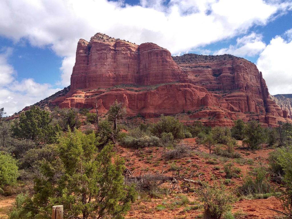 Gutsy Traveler: A large red rock formation rises above green shrubs and trees under a partly cloudy sky in Sedona, Arizona.