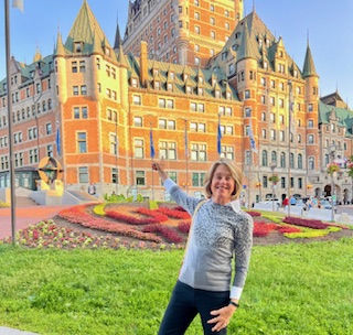 Gutsy Traveler: A woman stands on grass, smiling and pointing toward the historic Château Frontenac hotel in Quebec City—one of the top things to do in Quebec—with flowerbeds and the iconic building visible in the background.