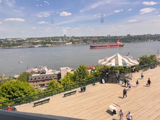 Gutsy Traveler: Enjoy a scenic view of Quebec City’s riverfront, where a large red ship passes by the boardwalk. Discover weekend guide highlights as people stroll past trees and a gazebo proudly flying the Canadian flag under a partly cloudy sky.