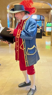 Gutsy Traveler: A man wearing a blue and red colonial-style costume with a large feathered hat holds a black folder while standing in a hotel lobby—a charming scene straight from any Quebec City weekend guide of things to do.
