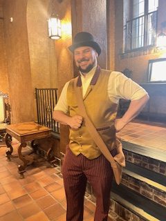 Gutsy Traveler: A man wearing a brown bowler hat, tan vest, and striped pants stands indoors on a tile floor, smiling with one hand on his hip—ready to explore Quebec City for your weekend guide of things to do.