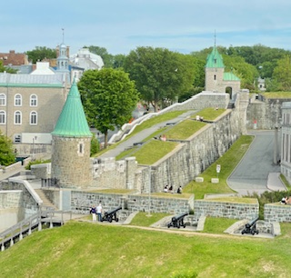 Gutsy Traveler: A historic stone fortress in Quebec City with green-roofed towers, sloped grassy walls, and cannons on display, surrounded by trees and nearby buildings—a must-see for any Weekend Guide of things to do.