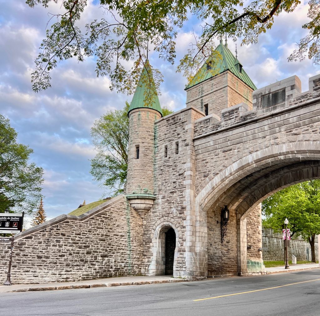 Gutsy Traveler: Stone archway with two green-roofed towers and a small doorway, set against a partly cloudy sky and surrounded by trees and a quiet street—one of Quebec City's best attractions and a must-see among things to do in the area.