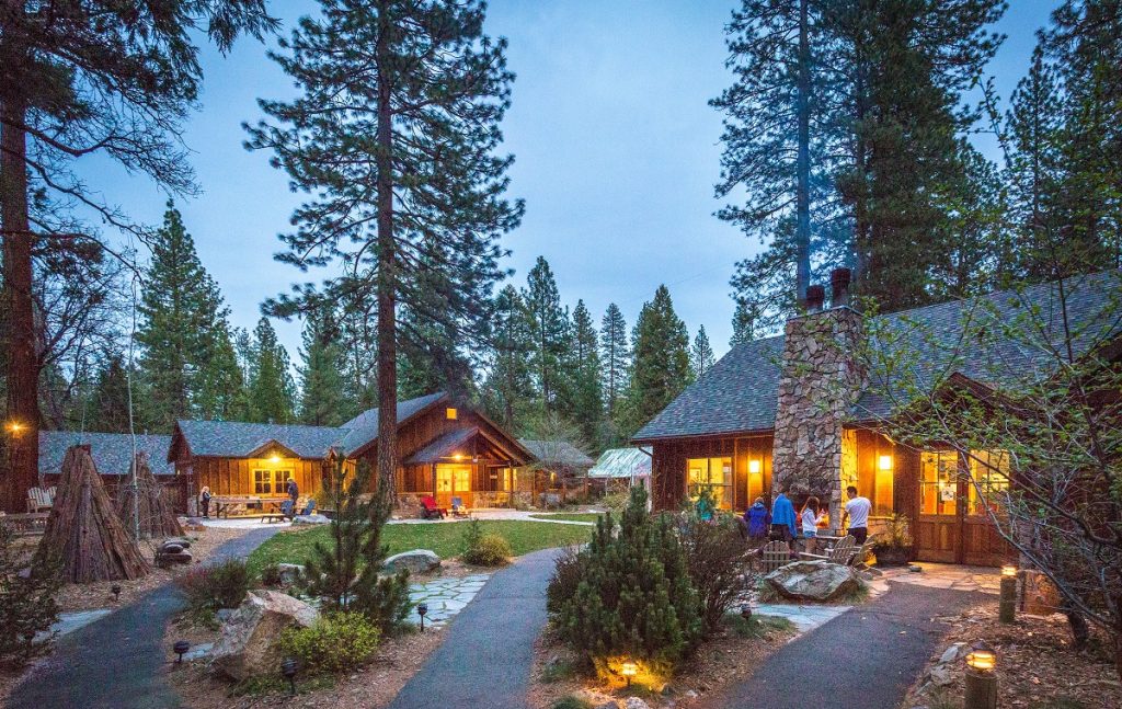 Gutsy Traveler: Several rustic cabins with lit windows are nestled among tall pine trees at dusk in Yosemite, with people gathered outside and a stone path winding through the landscaped grounds.