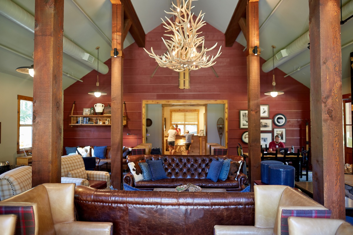 Gutsy Traveler: A rustic living room with leather sofas, armchairs, a large antler chandelier, wooden beams, and a red paneled wall captures the spirit of Yosemite during Off-Peak Hours, with people visible through a doorway in the background.