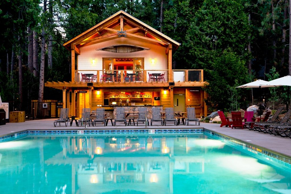 Gutsy Traveler: A wooden lodge with a balcony and outdoor seating overlooks a clear swimming pool, surrounded by lounge chairs, umbrellas, and forest trees—perfect for enjoying Yosemite tranquility during off-peak hours.
