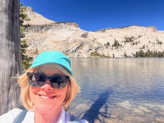 Gutsy Traveler: Woman wearing sunglasses and a teal cap takes a selfie by a clear mountain lake in Yosemite, with rocky hills and pine trees in the background, enjoying High Country Access under a clear blue sky.