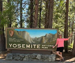Gutsy Traveler: A person in a pink shirt and black shorts stands next to a large "Yosemite National Park" sign, enjoying the scenic trees and peaceful atmosphere of Yosemite during off-peak seasons.