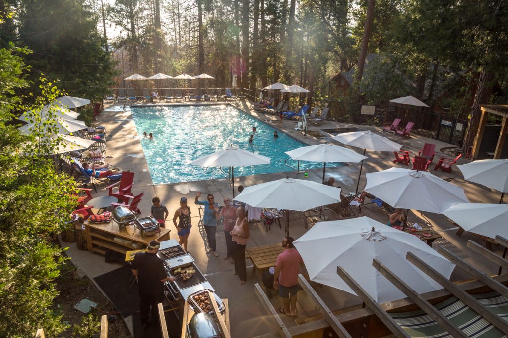 Gutsy Traveler: People relax and swim in an outdoor pool surrounded by lounge chairs and white umbrellas, with a group gathered near a barbecue area in a wooded setting—enjoying peaceful Yosemite vibes during off-peak hours.