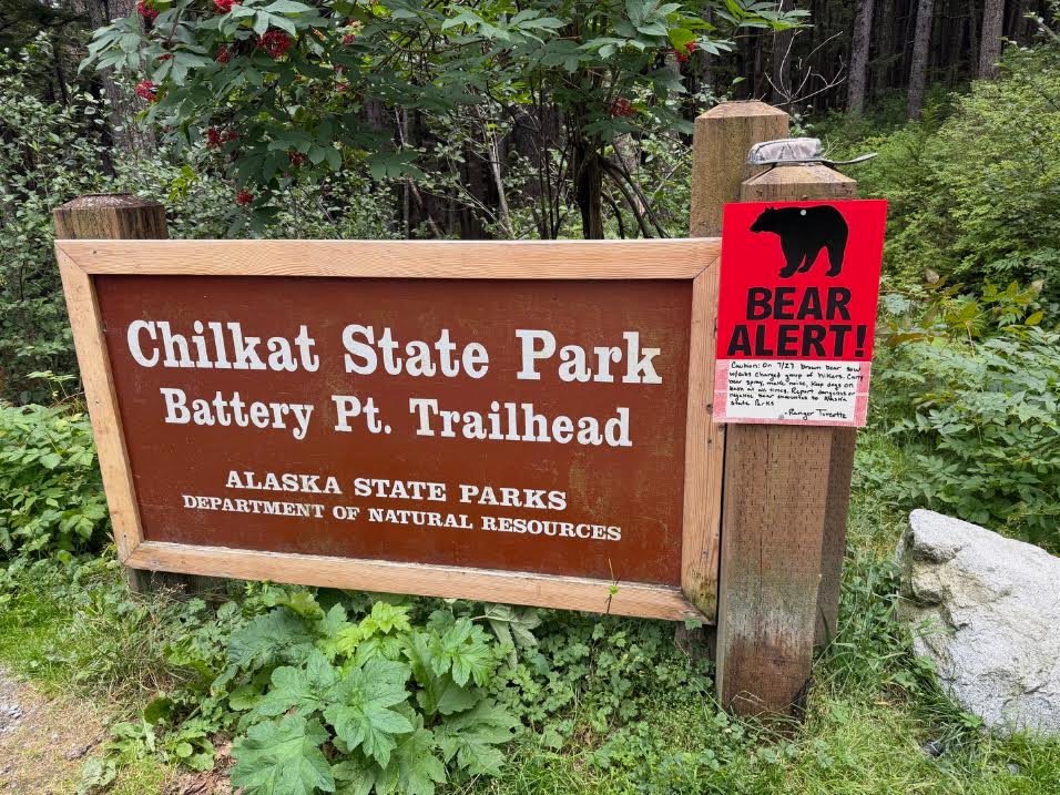 Gutsy Traveler: A wooden sign for the Chilkat State Park Battery Pt. Trailhead with a red bear alert sign attached, serving as a reminder of Alaska wildlife for those visiting the area.