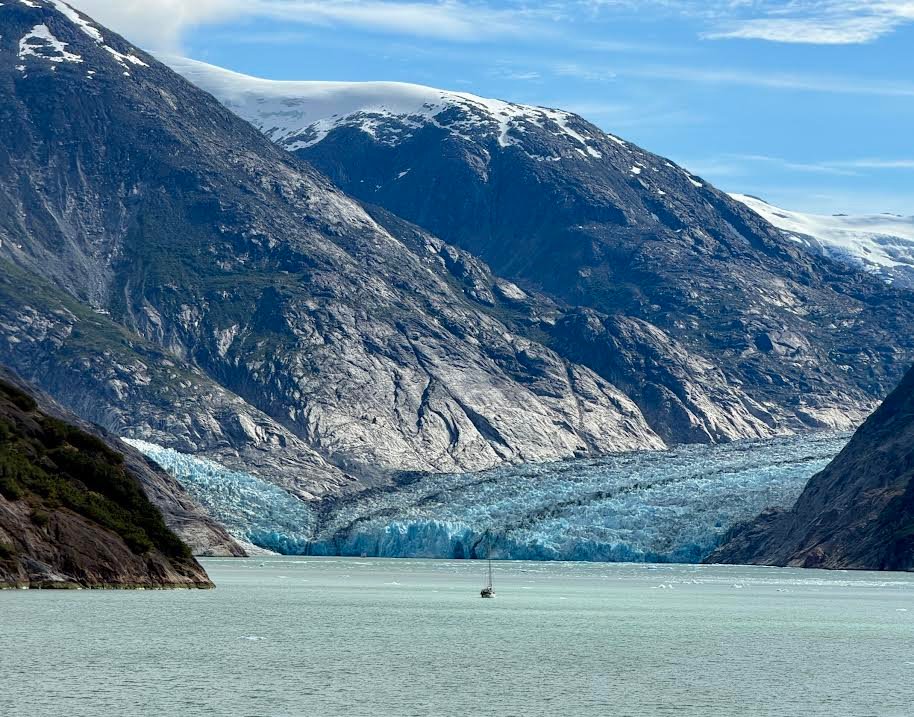 Gutsy Traveler: A small sailboat on the water in front of a blue glacier, nestled between snow-capped mountain peaks during an Alaska cruise.