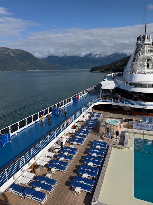 Gutsy Traveler: High-angle view of a cruise ship deck with rows of lounge chairs, a swimming pool, and a view of mountains across the water during an Alaska cruise.