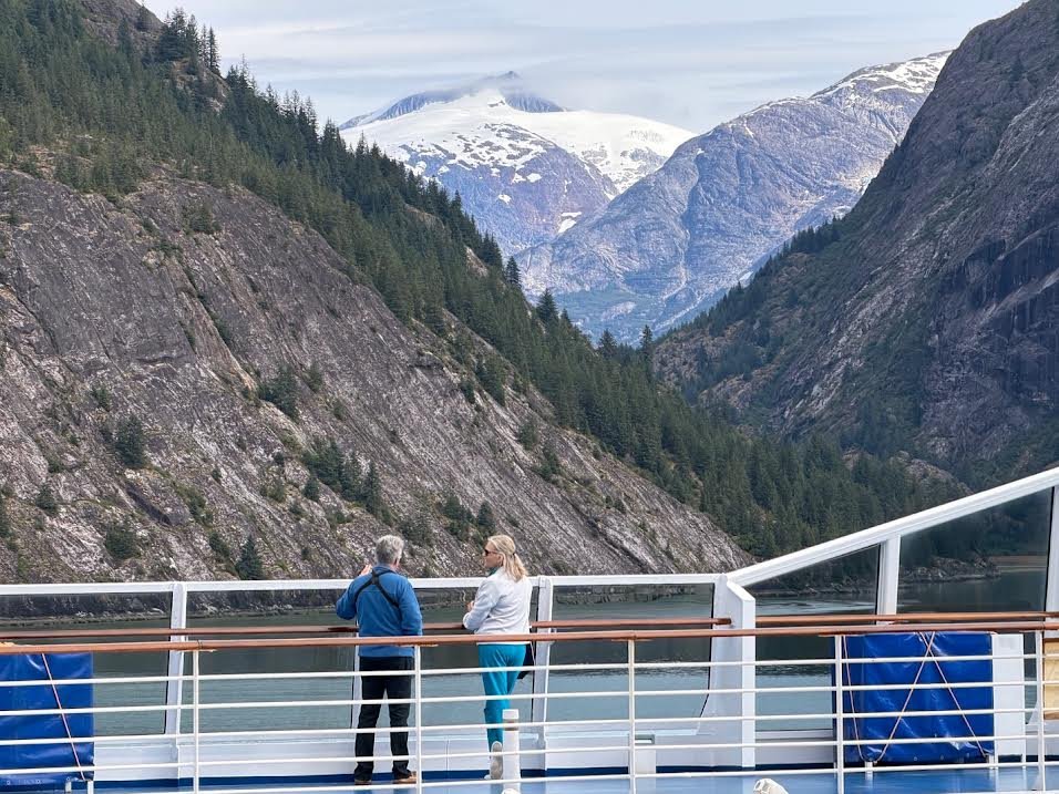 Gutsy Traveler: Two people stand on an Alaska cruise ship deck looking out at a fjord with steep, forested mountains and a snow-capped peak in the distance.