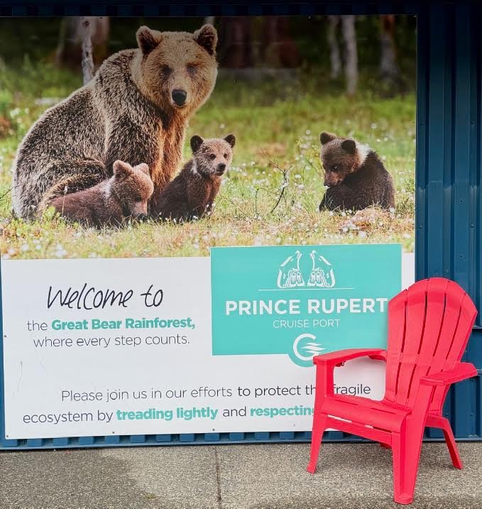 Gutsy Traveler: A red Adirondack chair sits in front of a Prince Rupert Cruise Port sign featuring a mother grizzly bear and her cubs, a common sight for Alaska travel.