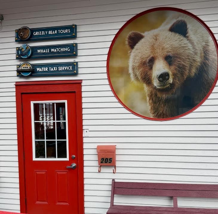Gutsy Traveler: Building exterior with signs for Alaska cruise excursions like Grizzly Bear Tours, whale watching, and water taxi service, positioned next to a circular grizzly bear mural and a red door.