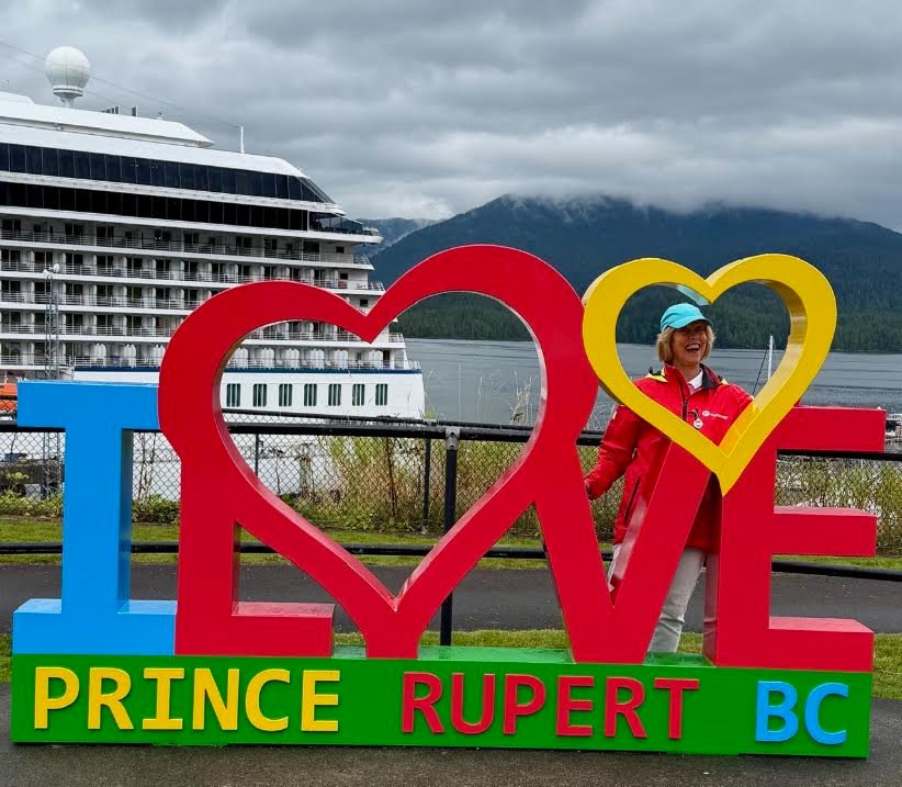 Gutsy Traveler: A person in a red jacket and turquoise cap poses inside the heart-shaped letter of a Prince Rupert sign, with an Alaska cruise ship docked in the background.