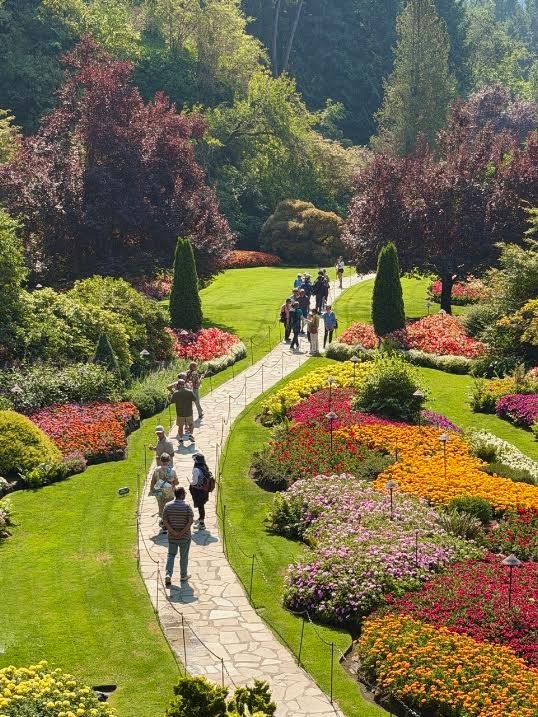 Gutsy Traveler: People walking along a stone path winding through a vibrant garden with colorful flower beds, a peaceful stop on an Alaska cruise.