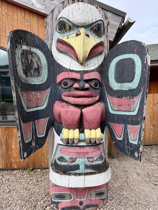 Gutsy Traveler: Carved wooden totem pole featuring a prominent eagle head at the top, a human-like figure in the center, and additional stylized animal motifs often seen during Alaska travel.