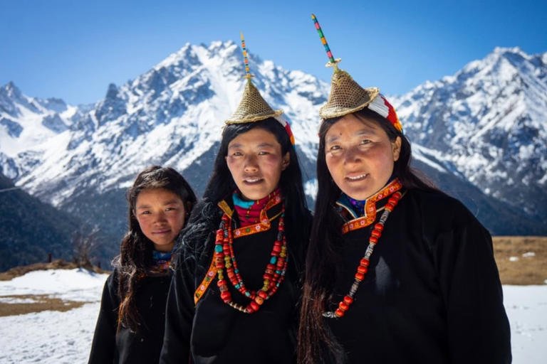 Gutsy Traveler: Three women in traditional black tunics, layered beaded necklaces, and pointed conical hats stand before snow-capped mountains in this Bhutan travel guide.